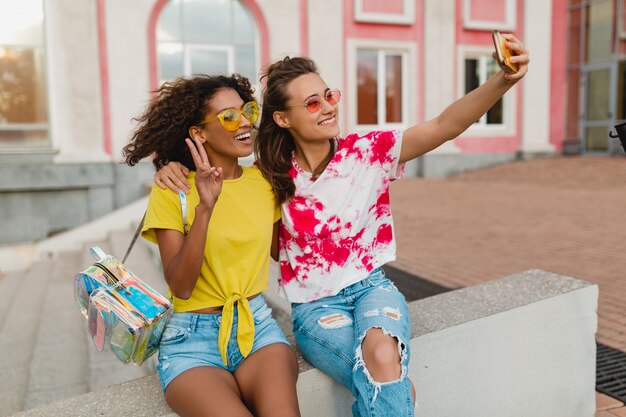 Счастливые молодые девушки друзья улыбаются, сидя на улице, принимая селфи фото на мобильный телефон, женщины веселятся вместе