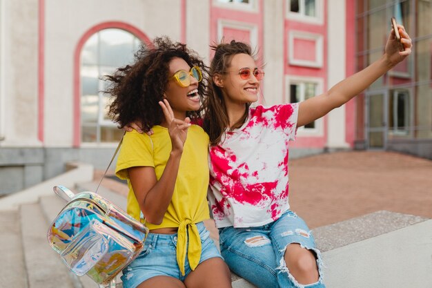 Happy young girls friends smiling sitting in street taking selfie photo on mobile phone, women having fun together