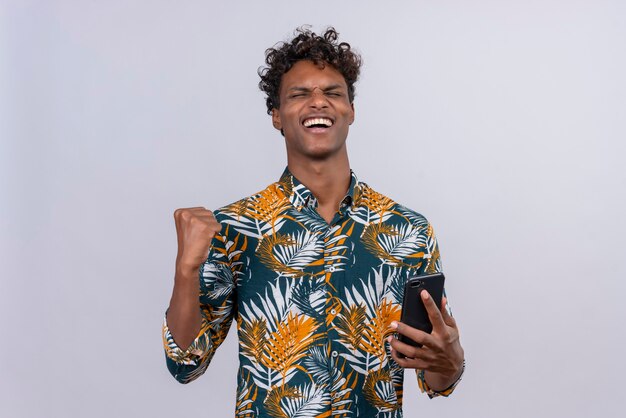 Happy young good-looking dark-skinned man with curly hair in leaves printed shirt holding his smartphone with clenching fist 