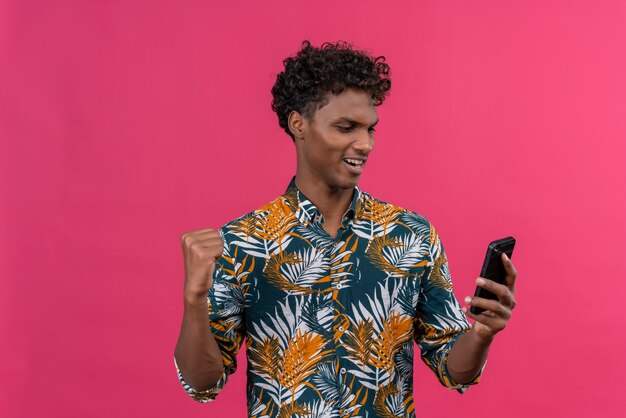 葉に巻き毛のある幸せな若いハンサムな黒肌の男性が拳を食いしばって彼のスマートフォンを見て葉のプリントシャツ