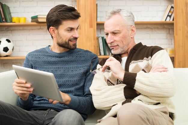 Happy young guy and aged man using tablet on settee