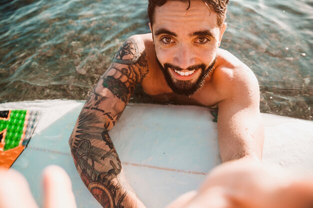 Happy young guy taking selfie and lying on surf board in water