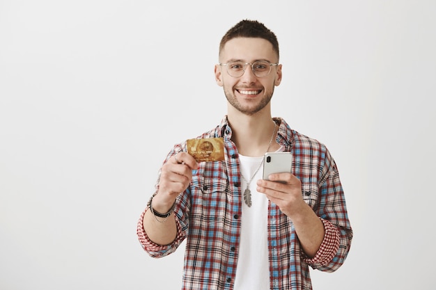 彼の携帯電話とカードでポーズをとって眼鏡をかけて幸せな若い男