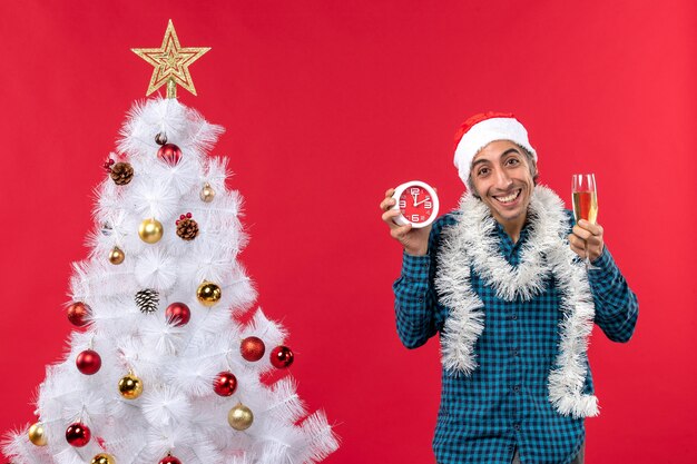 Happy young guy with santa claus hat and holding a glass of wine and clock standing near Christmas tree on red