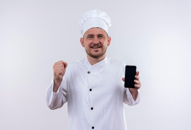 Happy young handsome cook in chef uniform holding mobile phone and raising fist isolated on white wall