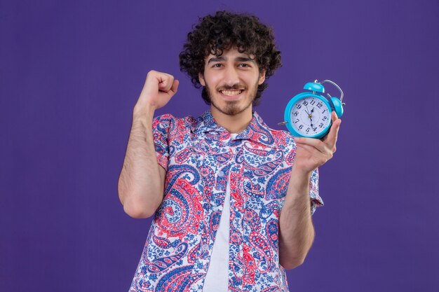 Happy young handsome curly traveler man holding alarm clock with raised fist on isolated purple space with copy space