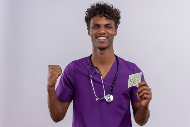 A happy young handsome dark-skinned male doctor with curly hair wearing violet uniform with stethoscope showing a paper card with the word yes 