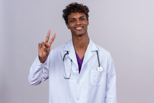 A happy young handsome dark-skinned male doctor with curly hair wearing white coat with stethoscope showing two fingers gesture 