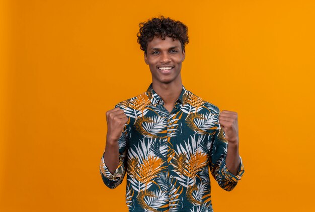 Happy young handsome dark-skinned man with curly hair in leaves printed shirt showing strength gesture with arms 