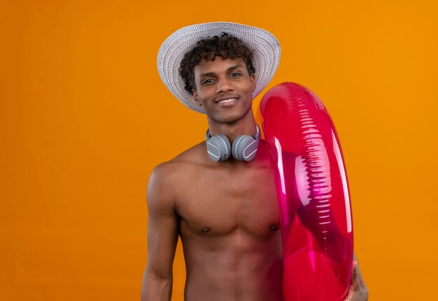 A happy young handsome dark-skinned man with curly hair wearing sun hat holding inflatable pool ring 
