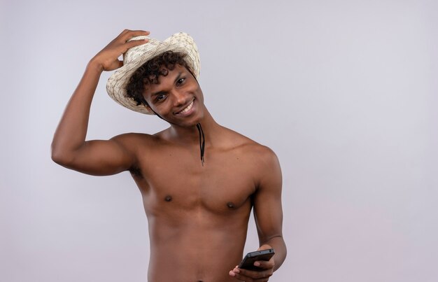 A happy young handsome dark-skinned man with curly hair wearing sun hat smiling and holding mobile phone 