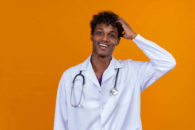 A happy young handsome dark-skinned man with curly hair wearing white coat with stethoscope  keeping hand on head 