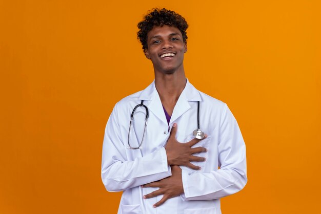 A happy young handsome dark-skinned man with curly hair wearing white coat with stethoscope smiling and holding hands 