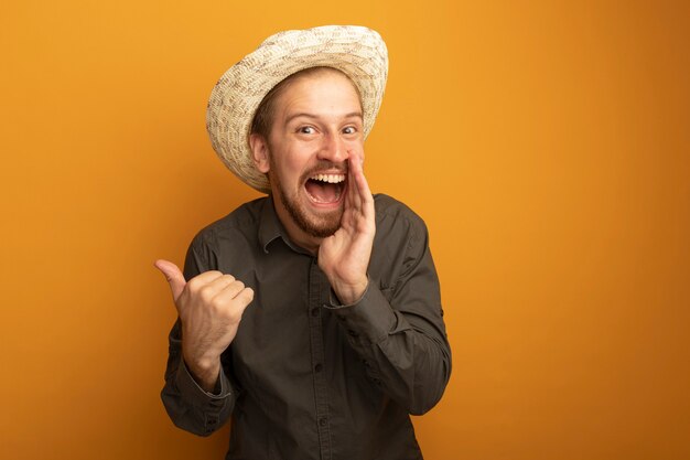 Happy young handsome man in grey shirt and summer hat shouting or calling someone with hand near mouth and pointing with index finger at something 