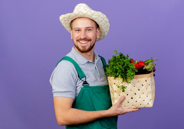 コピースペースと紫色の壁に分離された野菜のバスケットを保持している制服と帽子の幸せな若いハンサムなスラブ庭師