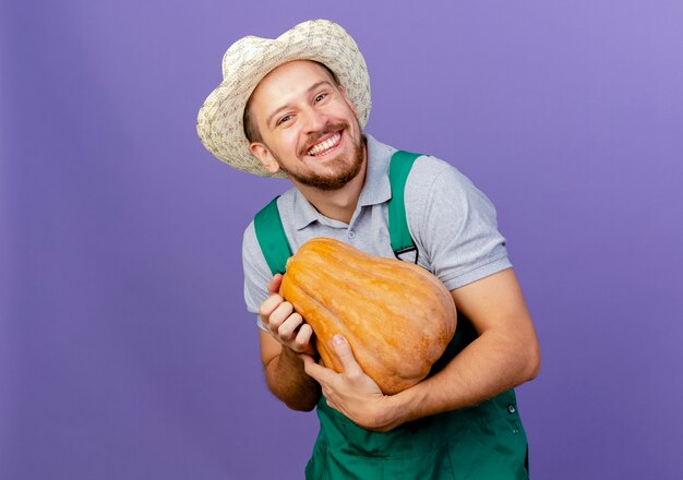 Happy young handsome slavic gardener in uniform and hat holding butternut pumpkin  isolated on purple wall with copy space