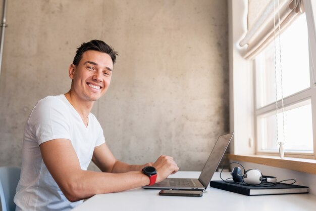 Happy young handsome smiling man in casual outfit sitting at table working on laptop