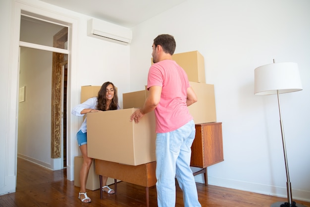 Happy young Hispanic couple moving into new flat, carrying carton boxes and furniture