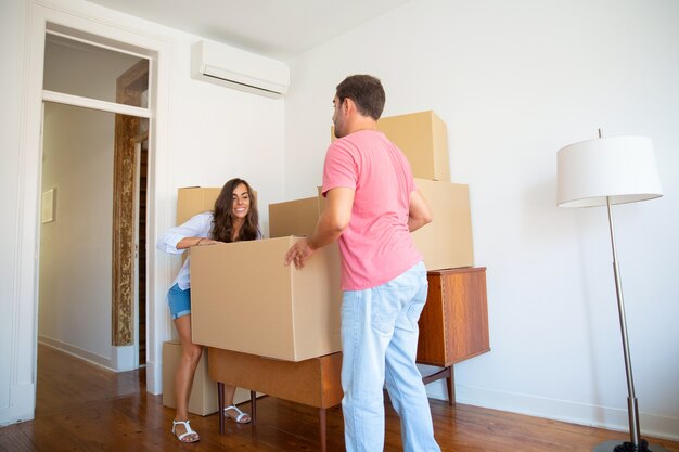 Happy young Hispanic couple moving into new flat, carrying carton boxes and furniture