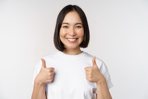 Happy young korean woman showing thumbs up and smiling give positive feedback recommending smth standing over white background