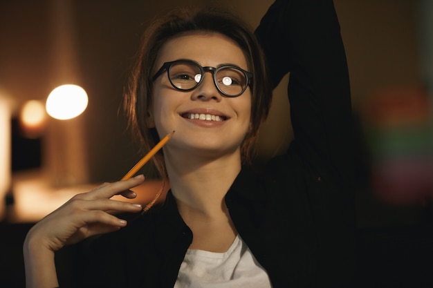 Счастливый молодой леди дизайнер, сидя в помещении ночью, держа карандаш