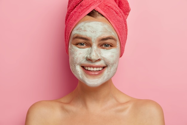 Happy young lady gets beauty mask on her beautiful face for softness, smiles broadly, stands topless indoor, cares about health and appearance, wrapped towel on head, expresses positive emotions.