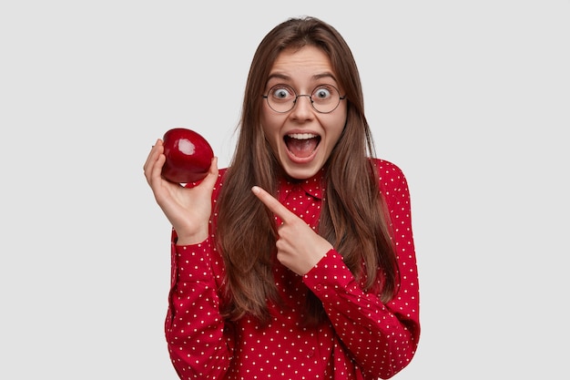 Happy young lady points at red juicy apple, demonstrates healthy food, keeps to diet, keeps jaw dropped, has long hair, amazed facial expression