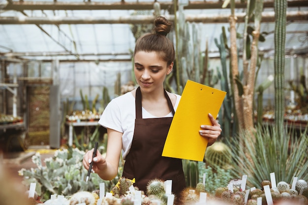 Happy young lady standing in greenhouse holding clipboard