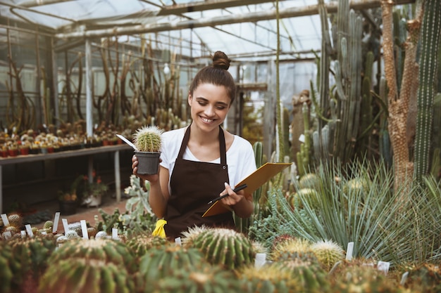 Happy young lady standing in greenhouse holding clipboard