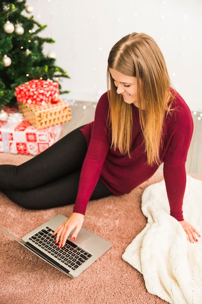 Happy young lady using laptop near gift boxes and Christmas tree