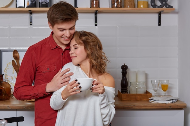 Happy young lovely couple on kitchen hugging each other. They enjoy spending time togehter. They drink coffee and smiling