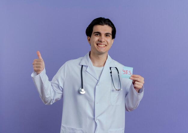 Счастливый молодой мужчина-врач в медицинском халате и стетоскопе показывает записку да, показывая большой палец вверх, изолированный на фиолетовой стене с копией пространства