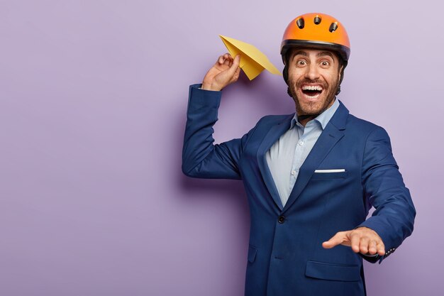 Happy young male engineer throws air plane, wears protective headgear and formal suit, has aim to develop his innovative project