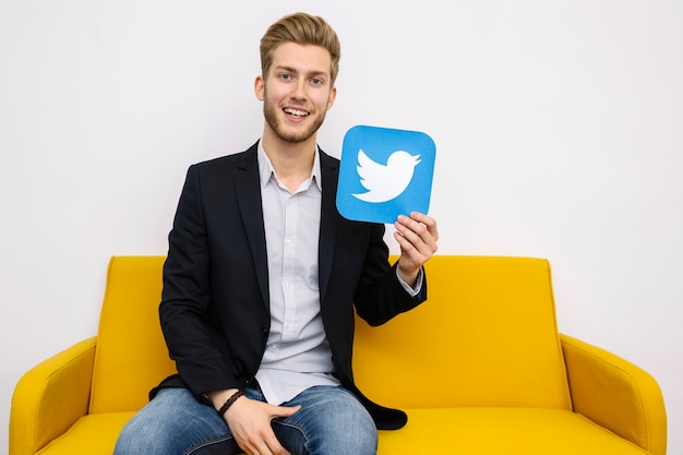 Happy young male sitting on yellow sofa holding twitter icon