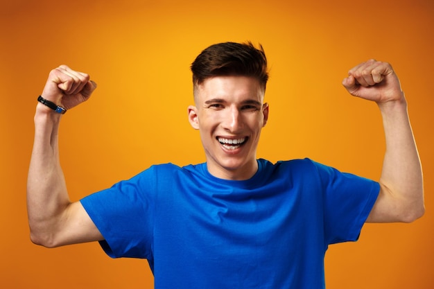 Счастливый молодой человек в синей футболке улыбается с поднятыми руками, празднуя успех