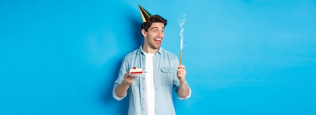 Foto gratuita giovane felice che festeggia il compleanno in cappello da festa che tiene la torta di compleanno e sorride in piedi sopra il blu b