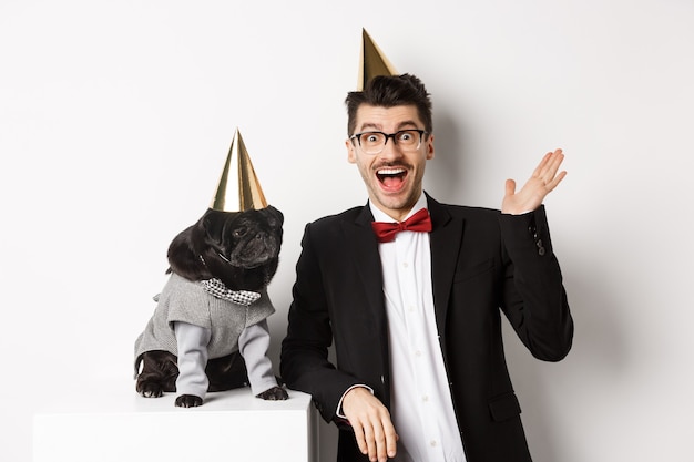 Happy young man and cute black dog wearing party cones, celebrating birthday, guy friendly saying hello and waving hand, standing over white background.