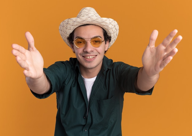 Happy young man in green shirt and summer hat wearing glasses looking at front with smile on face standing over orange wall