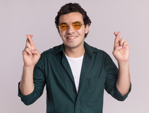 흰색 벽 위에 서있는 입술을 물고 바람직한 소원 횡단 손가락을 만드는 안경을 쓰고 녹색 셔츠에 행복 한 젊은 남자