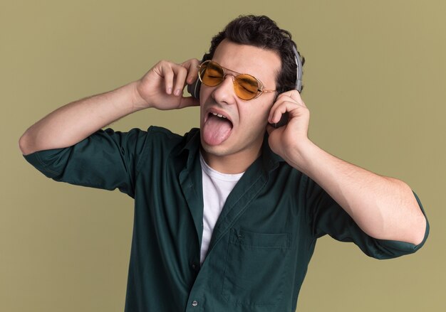 녹색 벽 위에 서 혀를 튀어 나와 그의 좋아하는 음악을 즐기는 헤드폰으로 안경을 쓰고 녹색 셔츠에 행복 한 젊은 남자