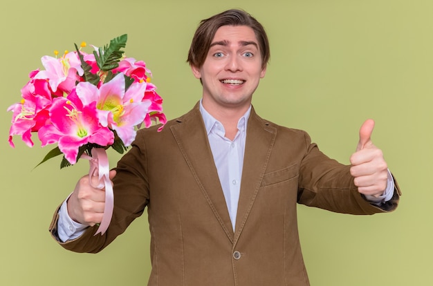 緑の壁の上に立っている国際女性の日を祝うために親指を上げて元気に笑って花の花束を持って幸せな若い男