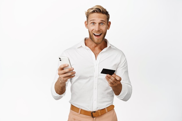 Happy young man holding mobile phone with credit card looking at camera surprised and smiling from good news white background