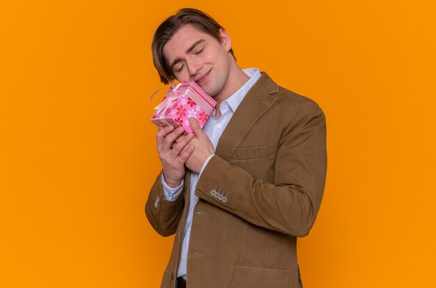 Happy young man holding present smiling cheerfully going to congratulate with international women's day standing over orange wall