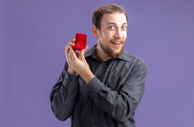 счастливый молодой человек держит красную коробку с обручальным кольцом, глядя в камеру, весело улыбаясь, концепция дня святого валентина, стоя над фиолетовой стеной