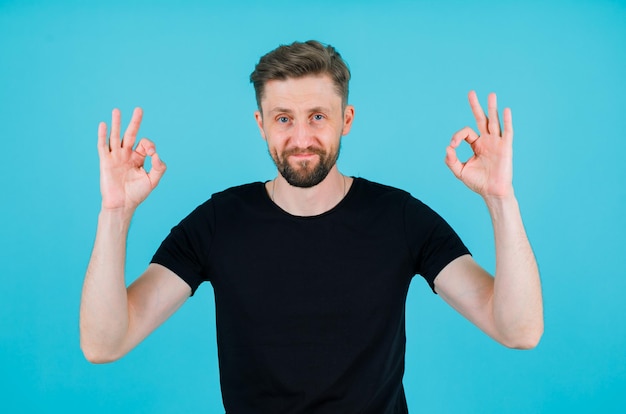 Happy young man is showing okay gestures by rasising up his hands on blue background