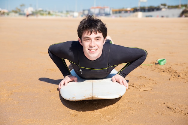 Happy young man lying on surfboard on sand