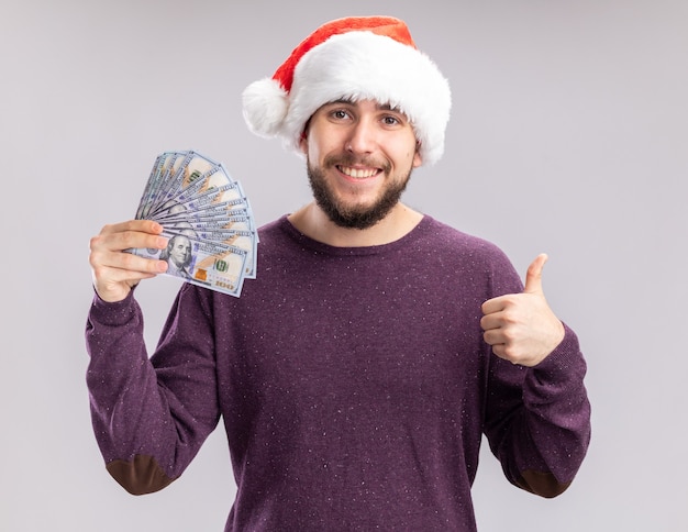 Felice giovane uomo in maglione viola e cappello santa tenendo contanti guardando la fotocamera sorridente che mostra i pollici in su in piedi su sfondo bianco