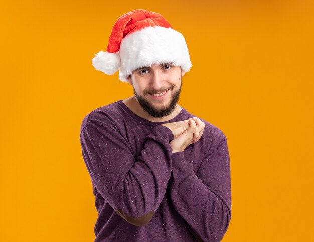 Счастливый молодой человек в фиолетовом свитере и шляпе санта-клауса, взявшись за руки вместе, смотрит в камеру с улыбкой на лице, стоя на оранжевом фоне