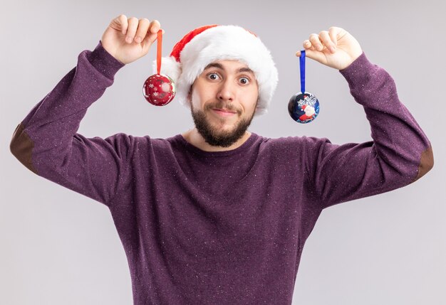 Happy young man in purple sweater and santa hat wearing funny glasses holding christmas balls looking at camera with smile on face standing over white background