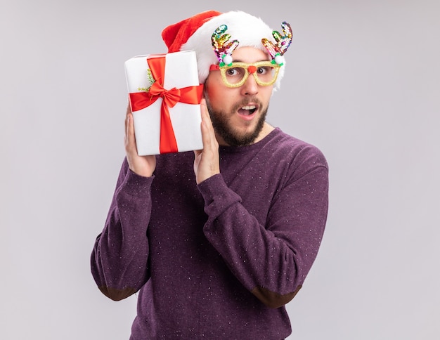 Happy young man in purple sweater and santa hat wearing funny glasses holding present looking at camera surprised , new year holiday concept standing over white background